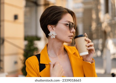 stylish beautiful elegant woman walking in city street wearing bright colorful yellow suit summer style, wearing sunglasses, drinking coffee, earrings accessories, short haircut haistyle
