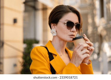 stylish beautiful elegant woman walking in city street wearing bright colorful yellow suit summer style, wearing sunglasses, drinking coffee, earrings accessories, short haircut haistyle