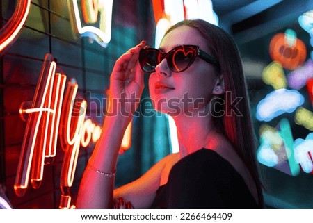 Stylish beautiful night model girl in black fashion dress wears sunglasses and walks on a dark background with neon signs