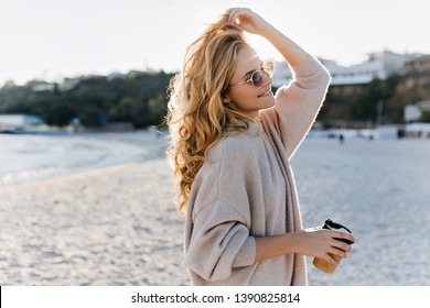 Stylish Beautiful Woman Blonde In Beige Oversized Sweater And Brown Sunglasses Walks Along Beach With Cardboard Cup Of Tea