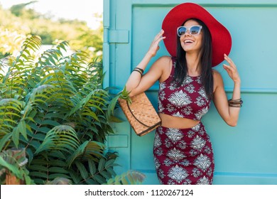 stylish beautiful woman in red hat posing on blue background, printed outfit, summer style, fashion trend, top, skirt, skinny, straw handbag, sunglasses, accessories, smiling, happy, tropical vacation