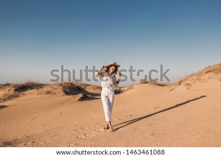 stylish beautiful woman walking in desert sand in white outfit wearing straw hat on sunset, travel safari on vacation