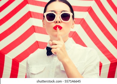 Stylish beautiful woman wearing white chemise and sunglasses with bright painted lips showing secret gesture next to a striped background