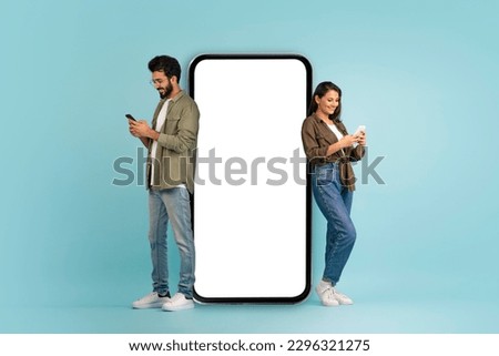 Stylish beautiful young eastern couple posing by huge phone with white blank screen, cheerful handsome indian man and pretty long-haired woman using smartphones, colorful background, mockup