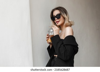 Stylish Beautiful Young Pretty Woman With Cool Modern Sunglasses In Elegant Black Clothes With Top And Blazer Drinking Cold Coffee With Juice And Walking On The Street  