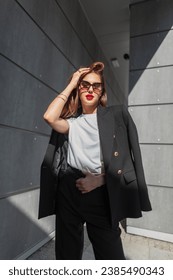Stylish beautiful young woman with vintage sunglasses with red lips in fashion urban casual clothes with a blazer and T-shirt stands near a gray building with sunlight. Pretty business girl