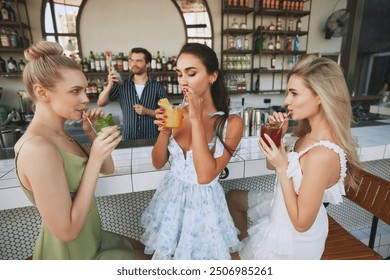 Stylish, beautiful young women in dresses enjoying drinks, cocktails in modern bar with bartender mixing drink on background. Chic bar atmosphere. Concept of friendship, party, leisure, celebration - Powered by Shutterstock