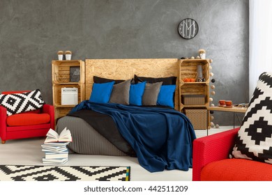 Stylish bedroom with grey walls and red armchair with little mess on bed. Wooden headboard of the bed and shelves made of wooden boxes