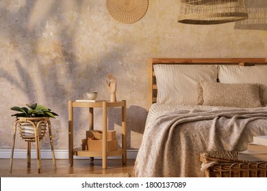 Stylish bedroom interior with design coffee table, furniture, plant, carpet, rattan decoration and elegant personal accessories. Beautiful beige bed sheets, blanket and pillows. Template.