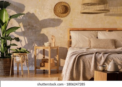 Stylish bedroom interior with design coffee table, furniture, tropical plant, rattan decoration and elegant personal accessories. Beautiful beige bed sheets, blanket and pillows. Template.