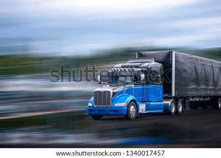 Stylish black and blue big rig long haul semi truck with aerodynamic spoilers transporting commercial cargo in full size black covered semi trailer moving on wide multiline divided highway road