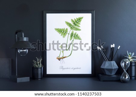 Stylish black desk with mock up photo frame, lamp,black accessories and plant. Black backgrounds wall. Design space in minimalistic interior.
