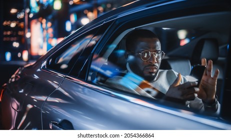 Stylish Black Man In Glasses Is Commuting Home In A Backseat Of A Taxi At Night. Handsome Male Using Smartphone And Looking Out Of Window While In A Car In Urban City Street With Working Neon Signs.