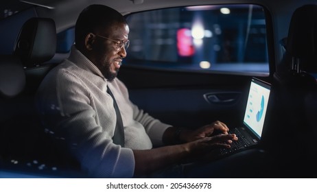 Stylish Black Man In Glasses Is Commuting Home In A Backseat Of A Taxi At Night. Male Using Laptop Computer And Looking Out Of Window While In A Car In Urban City Street With Working Neon Signs.