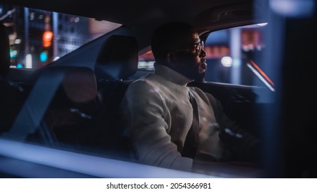 Stylish Black Man In Glasses Is Commuting Home In A Backseat Of A Taxi At Night. Handsome Male Passenger Looking Out Of Window While In A Car In Urban City Street With Working Neon Signs.