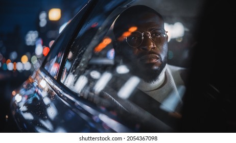 Stylish Black Man In Glasses Is Commuting Home In A Backseat Of A Taxi On A Rainy Night. Handsome Male Passenger Looking Out Of Window While In A Car In Urban City Street With Working Neon Signs.
