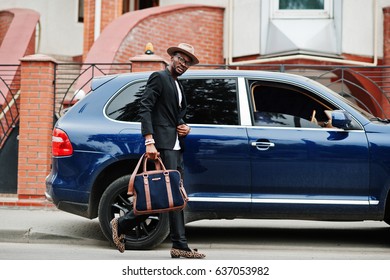 Stylish Black Man At Glasses With Hat, Wear On Suit With Handbag Against Luxury Car. Rich African American Businessman.