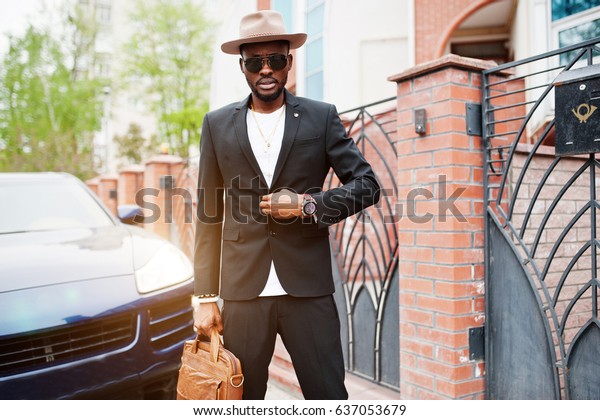 Stylish\
black man at glasses with hat, wear on suit with handbag against\
luxury car. Rich african american\
businessman.