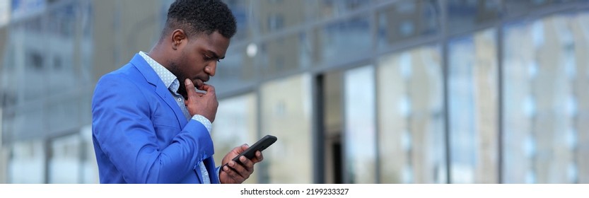 Stylish Black Man Looks At Smartphone With Thoughtful Expression Against Blurry Office Building. African American Ceo Thinks About Future Plans