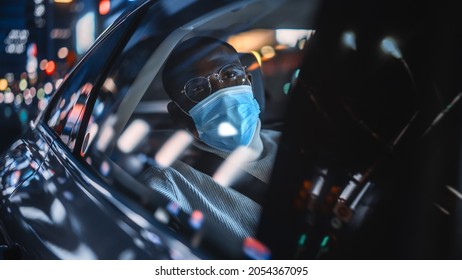 Stylish Black Man Wearing A Face Mask Commuting Home In A Backseat Of A Taxi At Night. Handsome Male Passenger Looking Out Of Window While In A Car In Urban City Street With Working Neon Signs.