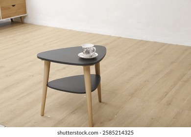 Stylish black triangular coffee table with a lower shelf, topped with a tea cup, perfect for enhancing modern home interiors.