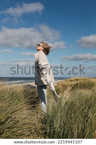 Stylish blonde girl standing alone at the winter time sea beach on the wind, smiling with her arms wide spread enjoying the sun. Idea of freedom, power, nature, solitude. Cold sea shore with grass.