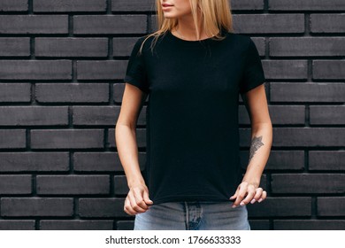 Stylish blonde girl wearing black t-shirt and glasses posing on black wall background, urban clothing style. Street photography