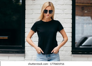 Stylish blonde girl wearing black t-shirt and glasses posing against street , urban clothing style. Street photography