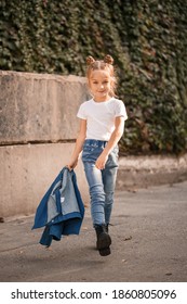 Stylish Blonde Little Girl In Jeans And A White T-shirt Walks Down The Street. Girl 7 Years Old Small Model, Beautiful Child