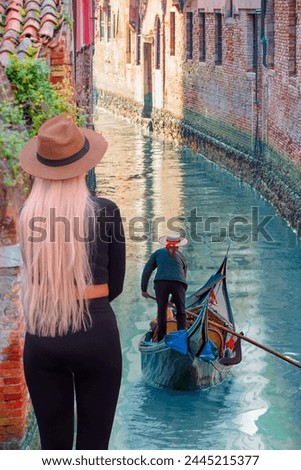 A stylish blonde woman on the bridge watching the Venetian gondolier - Venetian gondolier punting gondola through green canal waters of Venice Italy