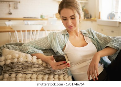 Stylish blonde young woman sitting on couch with smart phone, messaging friends online. Beautiful female teenager checking newsfeed via her social media account, typing comments, liking pics