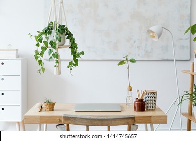 Stylish and boho home interior of open work space with wooden desk, chair, lamp, laptop and white shelf. Design and elegant personal accessories. Botany and minimalistic home decor with plants.