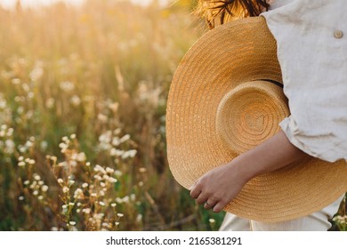 Stylish boho woman walking with straw hat in hand close up among wildflowers in sunset light. Atmospheric moment. Summer travel. Young female in rustic linen cloth relaxing in summer meadow - Powered by Shutterstock