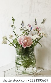 Stylish bouquet with peony and wildflowers on tile shelf on rustic wall background in modern room. Beautiful flowers in glass vase gathered from garden, summer floral arrangement in home. - Shutterstock ID 2321395637