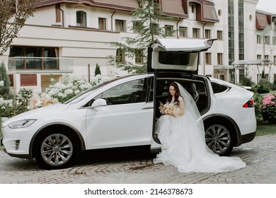 Stylish Bride with Flower Bouquet near Limousine. Pretty Young Woman Dressed White Wedding Dress and Jewelry Diadem with Veil. Girl Holding Roses Bunch and Standing near white car. Outside Photo