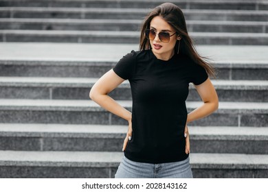Stylish brunette girl wearing black t-shirt and glasses posing against street , urban clothing style. Street photography