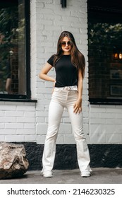 Stylish brunette girl wearing black t-shirt and white flared pants posing against street , urban clothing style. Street photography