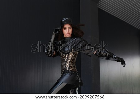 Stylish brunette model with vivid makeup, gloves, corset with leopard pattern, Panama hat posing over black wall urban building. Wearing total black leather clothes. Latex contemporary fashion.