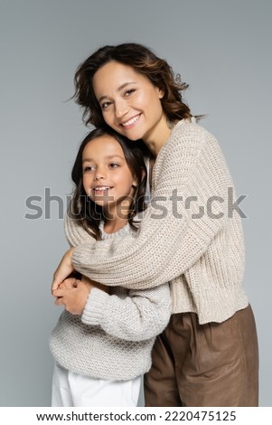 stylish brunette woman looking at camera while hugging daughter isolated on grey