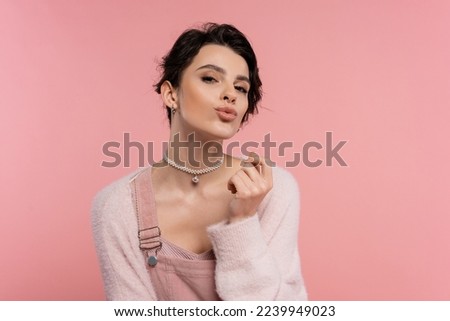 stylish brunette woman in pearl necklace pouting lips and looking at camera isolated on pink