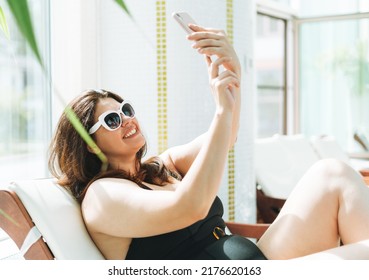 Stylish brunette woman plus size body positive in black swimsuit and sunglasses taking selfie on mobile phone on lounger in spa - Powered by Shutterstock