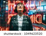 Stylish brunette woman in trendy apparel and eyewear looking up enjoying nightlife in city. Gorgeous fashion hipster girl in leather jacket standing outdoors on street with neon city illumination