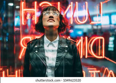Stylish brunette woman in trendy apparel and eyewear looking up enjoying nightlife in city. Gorgeous fashion hipster girl in leather jacket standing outdoors on street with neon city illumination