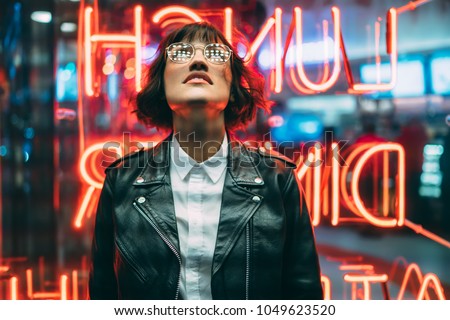 Stylish brunette woman in trendy apparel and eyewear looking up enjoying nightlife in city. Gorgeous fashion hipster girl in leather jacket standing outdoors on street with neon city illumination
