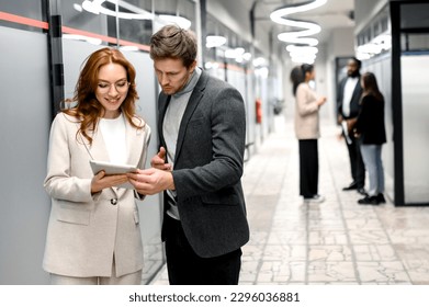 Stylish business people are standing in office, using a tablet. Handsome male employee asking an advice from beautiful redhead woman colleague about work questions - Powered by Shutterstock