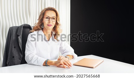 Stylish business woman at a work desk. A successful professional leader. Beautiful woman in a white shirt, glasses and jewelry in an office chair.