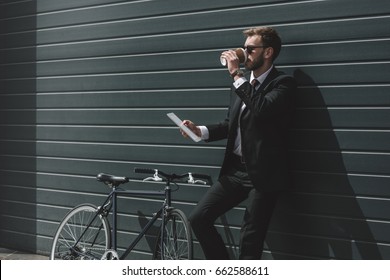 stylish businessman with bicycle using digital tablet and drinking coffee while standing in front of black wall - Powered by Shutterstock