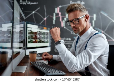 Stylish businessman, trader in glasses sitting in front of computer monitor while working using wireless earphones. Blackboard full of data analyses in the background. Stock trading, people concept.