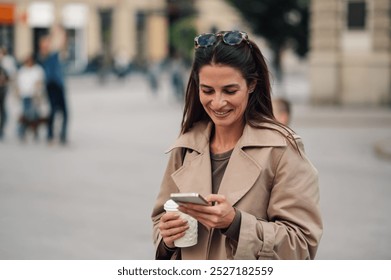 Stylish businesswoman multitasks on city streets, using her smartphone and holding coffee, embodying modern urban life with a cheerful, positive energy - Powered by Shutterstock