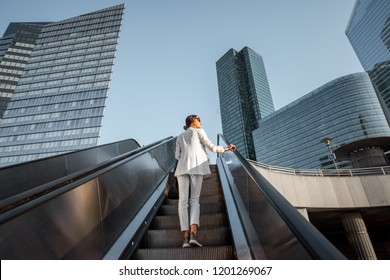 Stylish businesswoman in white suit going up on the escalator at the business centre outdoors with skyscrapers on the background in Paris